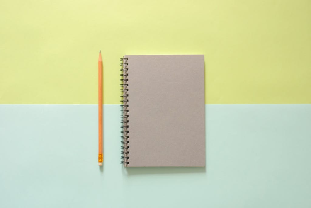 Flat lay of a spiral notebook and pencil on a pastel background, ideal for school and office themes.