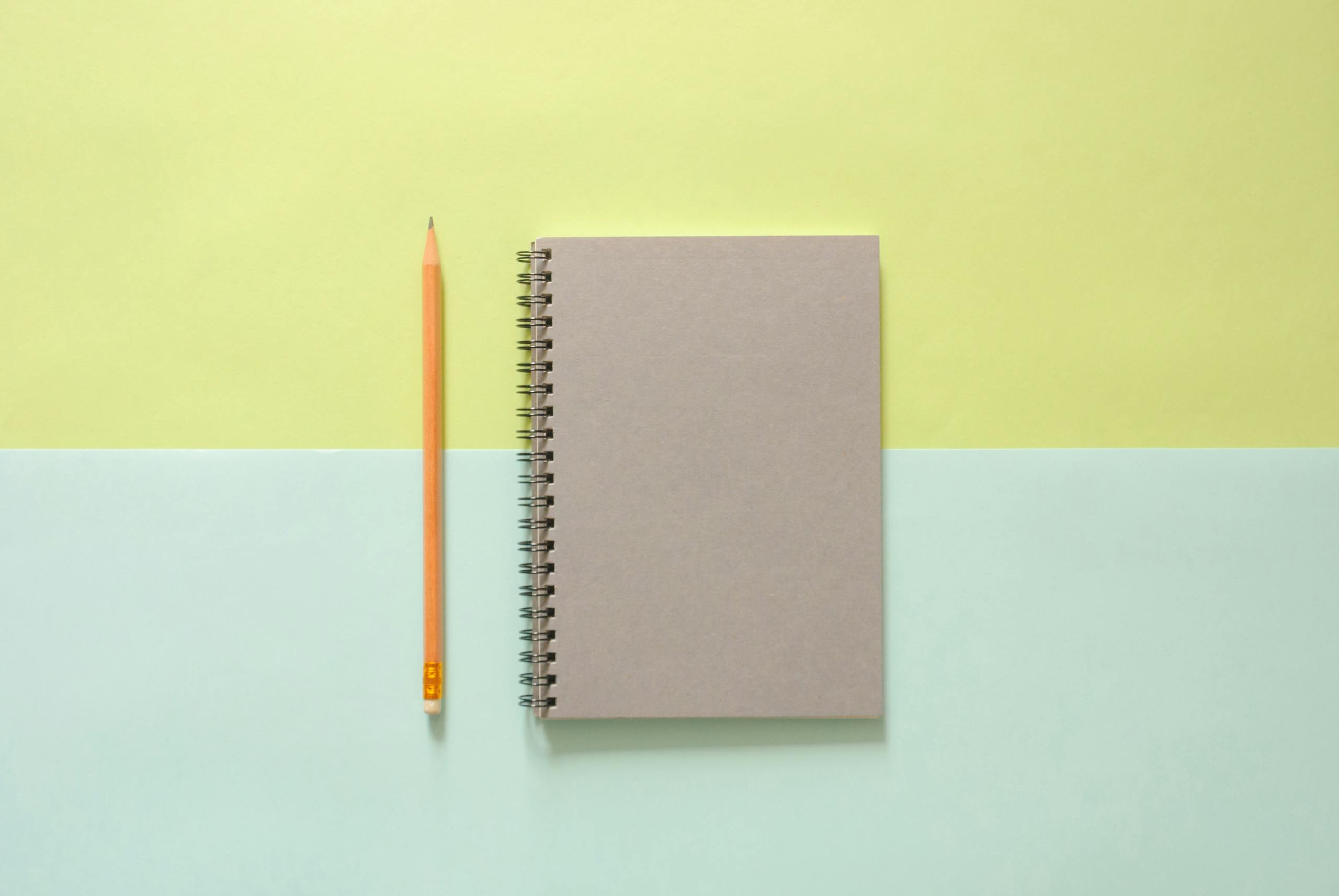 Flat lay of a spiral notebook and pencil on a pastel background, ideal for school and office themes.